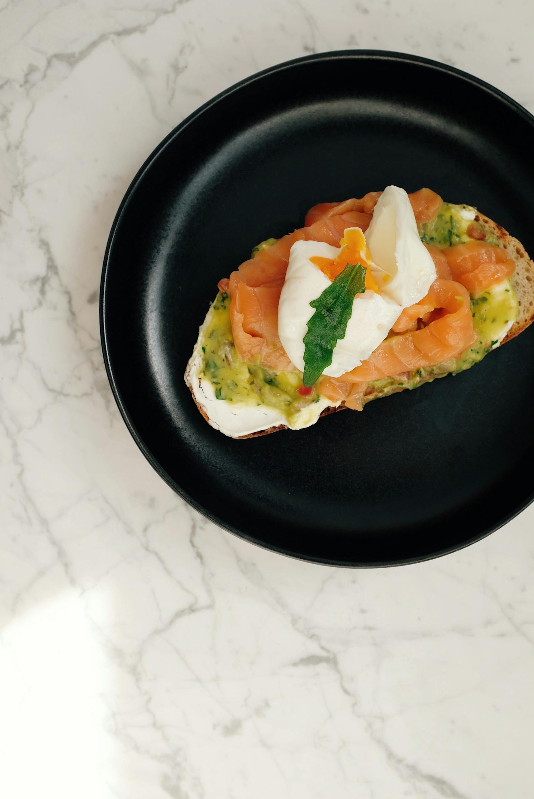 Delicious poached egg with salmon on toast, garnished with greens, served on a black plate.